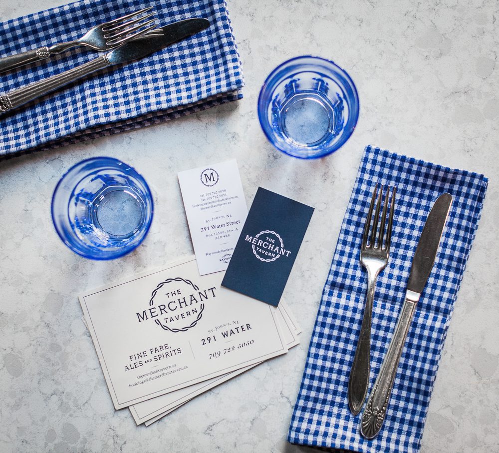 An overhead view of blue drinking glasses, cutlery placesettings and Merchant Tavern postcards and business cards on a table
